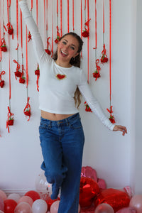 Woman in a white sweater and blue jeans standing in front of a decorative wall with red ribbons and balloons.