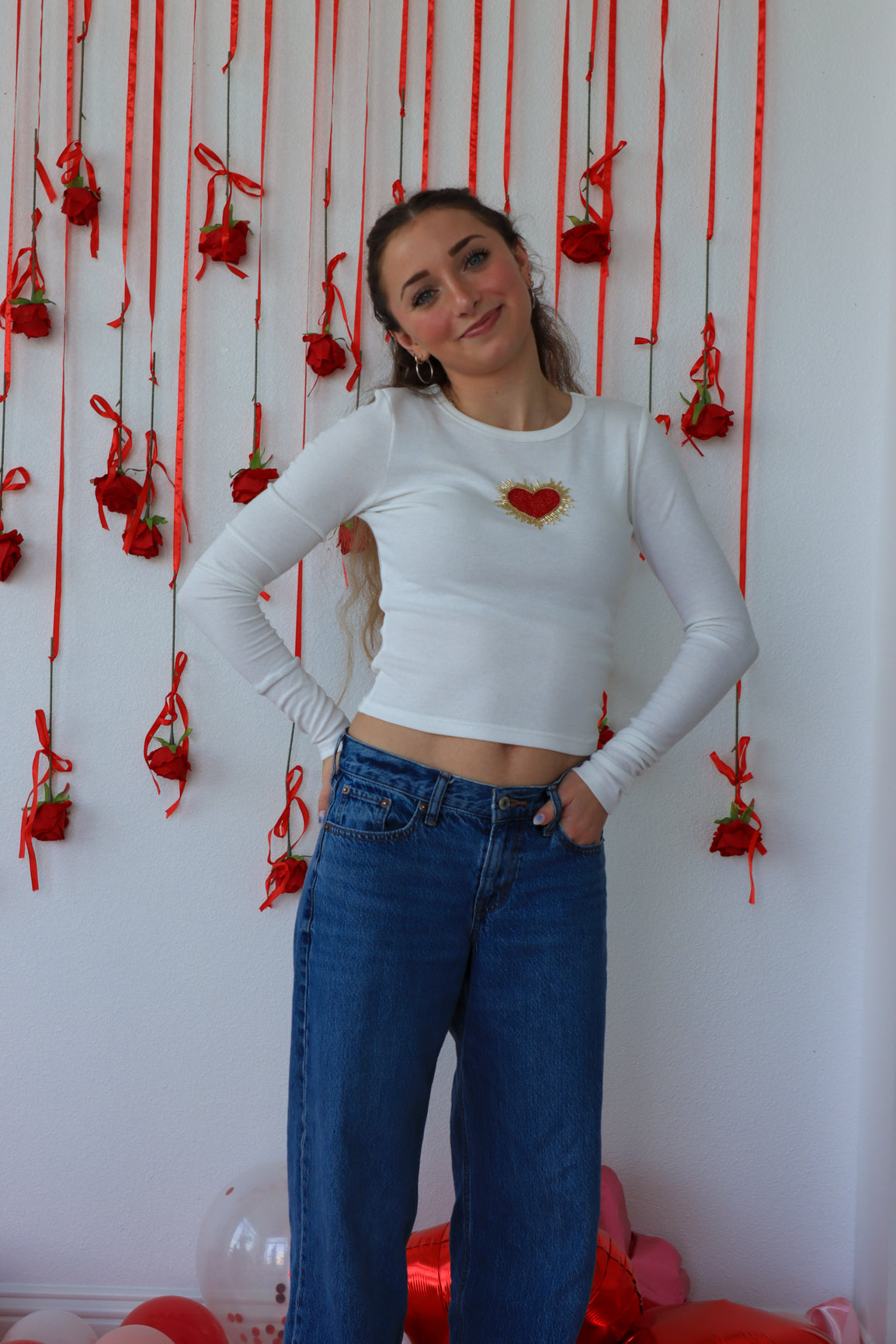 Woman wearing a white long-sleeve top with a red heart design and blue jeans standing against a wall with red decorations.