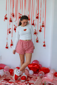 Person in a white long-sleeve top and red checkered shorts standing in a room decorated with red and pink balloons and streamers.