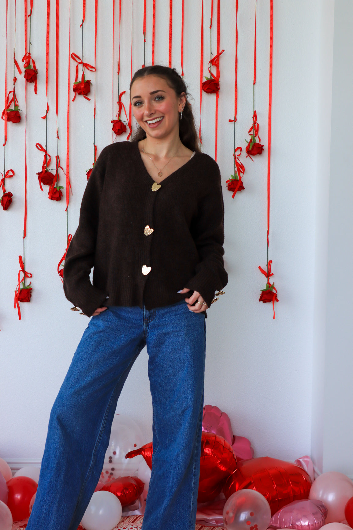 Woman in a black cardigan and blue jeans standing in front of a decorative wall with red ribbons and balloons.