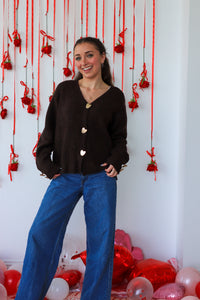 Woman in a black cardigan and blue jeans standing in front of a decorative wall with red ribbons and balloons.