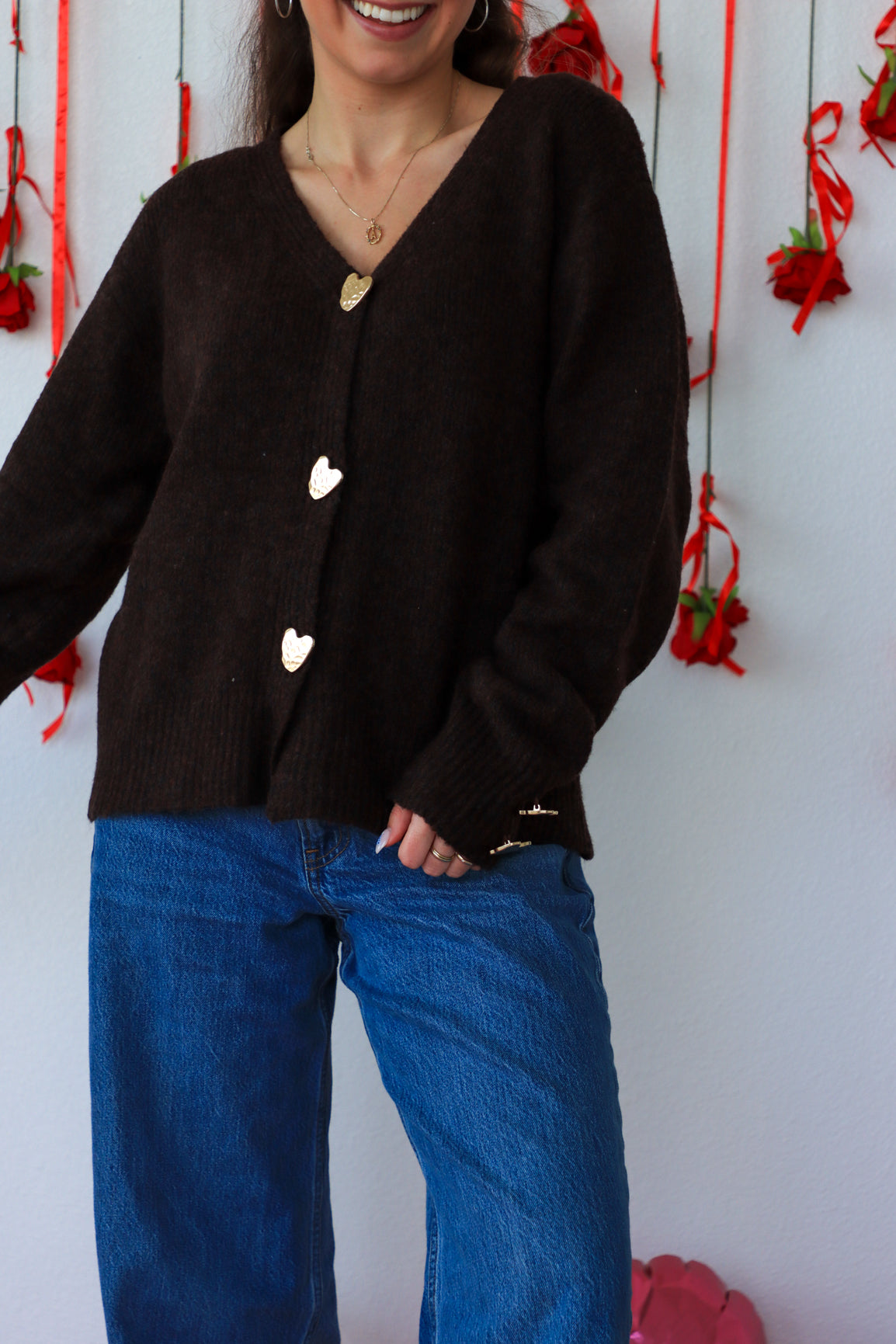 Person wearing a black cardigan with heart buttons and blue jeans against a white wall with red decorations.