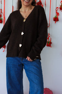 Person wearing a black cardigan with heart buttons and blue jeans against a white wall with red decorations.