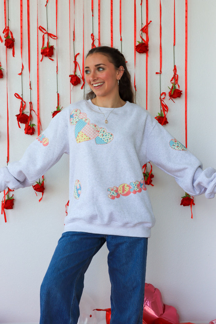 Person wearing a light gray sweatshirt with colorful patterns in front of a decorative wall with red ribbons and flowers.