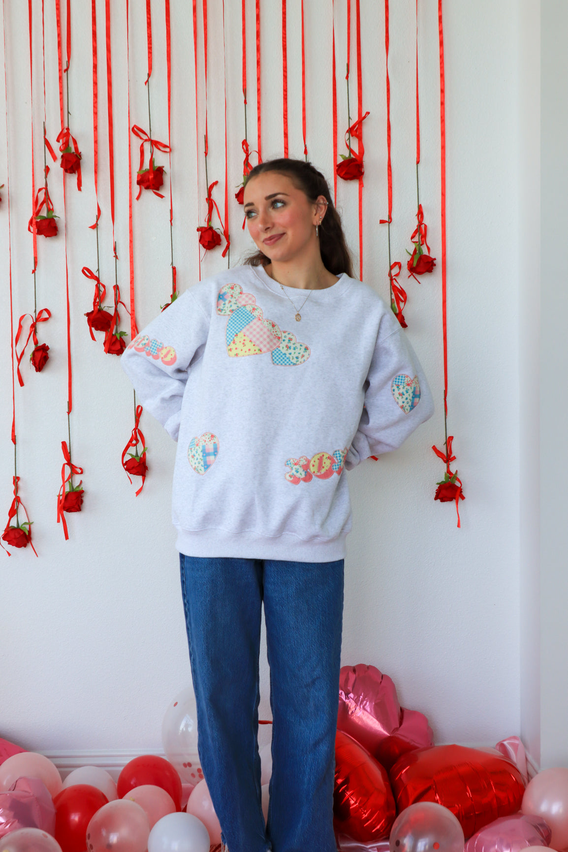 Woman wearing a colorful sweater with heart designs in front of a decorative wall with red ribbons and balloons.