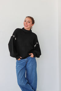 Person wearing a black sweater with white patterns and blue jeans against a plain background