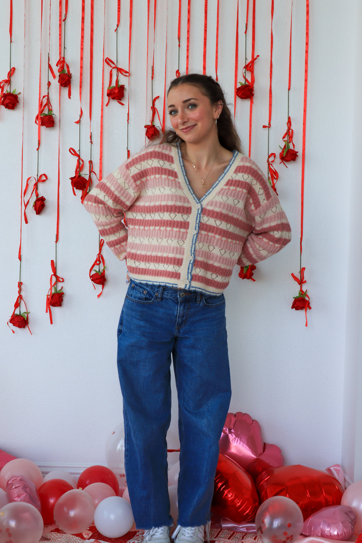 Woman wearing a striped sweater and blue jeans standing against a wall with red decorations and balloons on the floor.