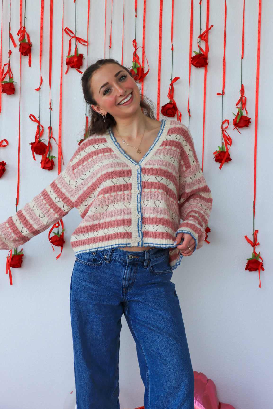 Woman wearing a pink and white striped sweater with blue jeans standing against a wall with red decorations.