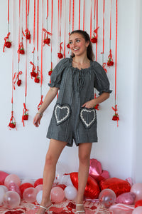 Woman in a gray dress with heart patterns standing in front of a decorative wall with red ribbons and balloons.