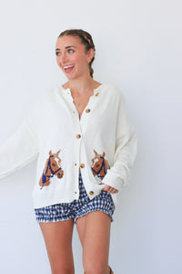 Woman wearing a white cardigan with horse designs and blue checkered shorts against a white background