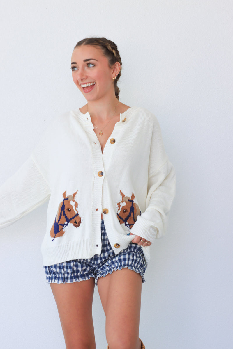 Woman wearing a white cardigan with horse designs and blue checkered shorts against a white background