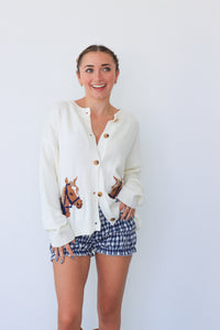 Woman wearing a white cardigan with horse designs and plaid shorts on a light blue background
