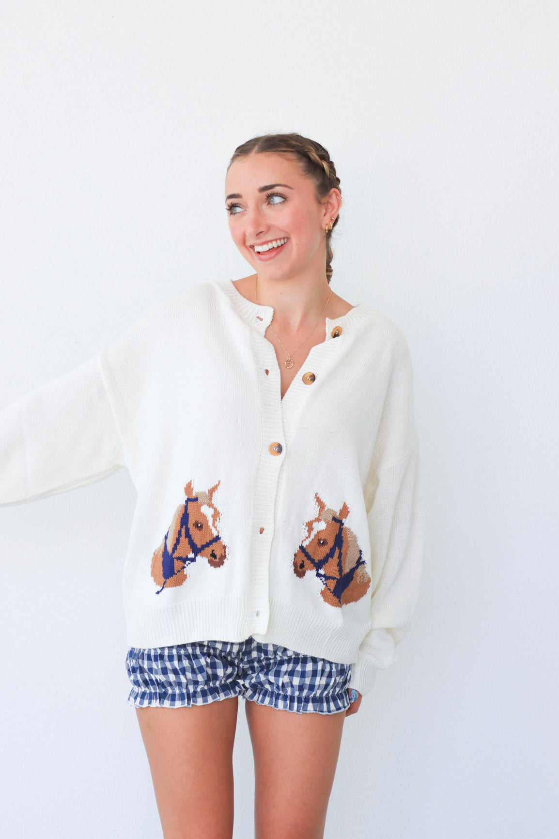 Woman wearing a white cardigan with horse designs and blue checkered shorts on a white background