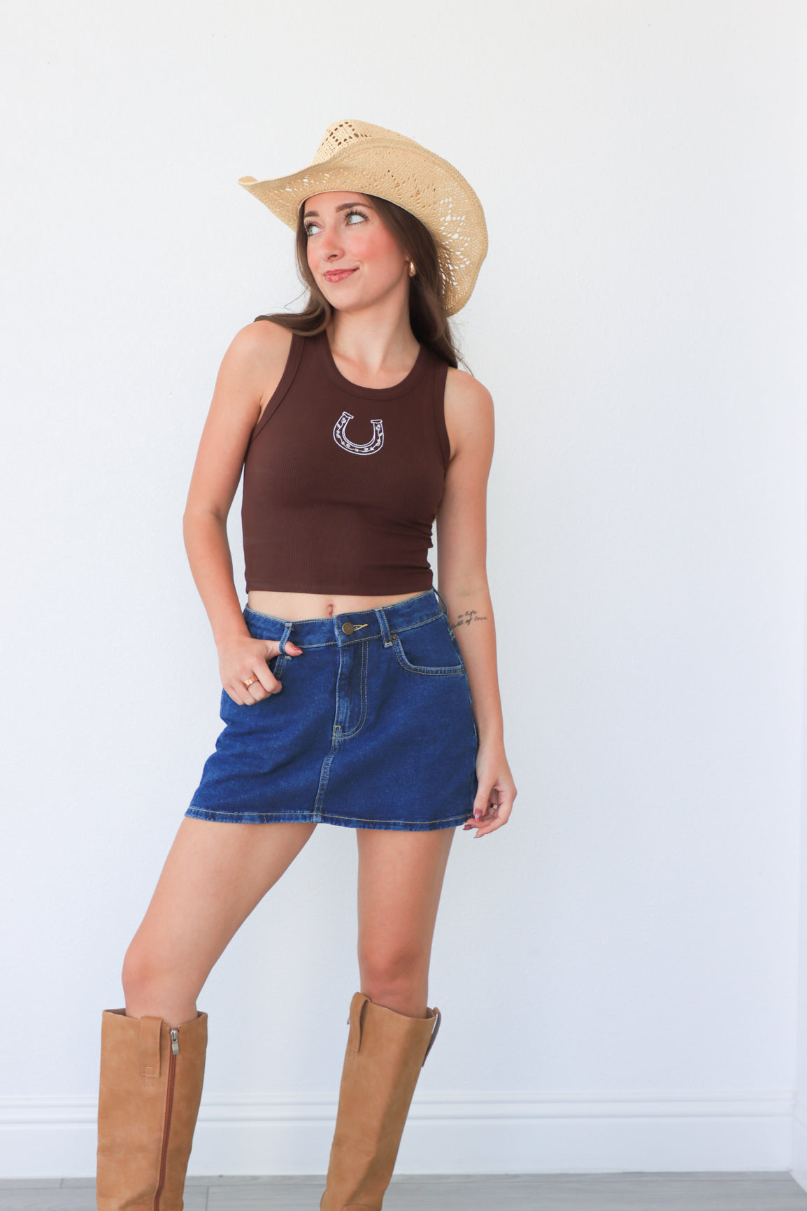 Woman wearing a brown tank top with a horseshoe design, blue denim shorts, and tan cowboy boots against a white background.