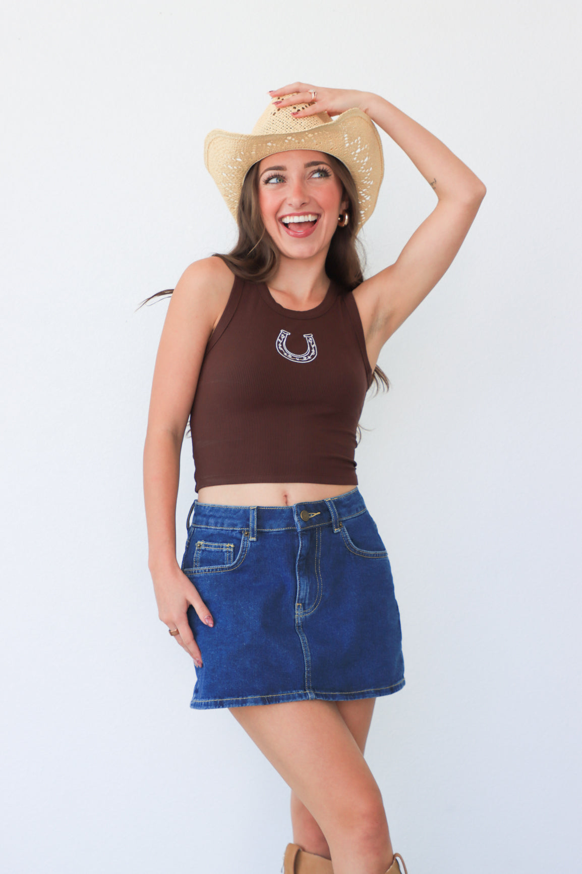 Woman wearing a brown tank top with a horse shoe design, blue denim skirt, and cowboy hat on a white background