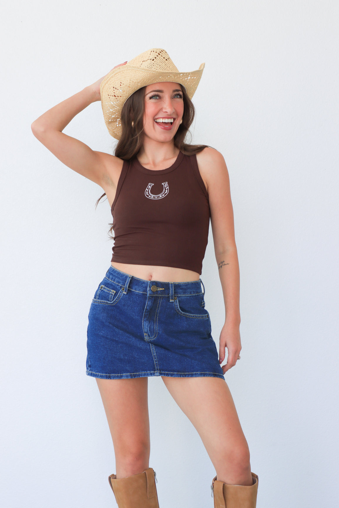 Woman wearing a brown tank top with a horse shoe design, blue denim shorts, and a straw cowboy hat on a white background.