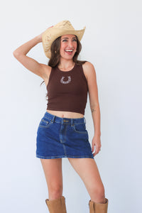 Woman wearing a brown tank top with a horse shoe design, blue denim shorts, and a straw cowboy hat on a white background.