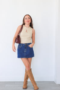 Woman wearing a beige sleeveless top, blue denim skirt, and tan knee-high boots against a white background.