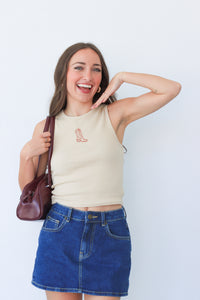 Woman wearing a beige tank top and blue denim skirt, holding a brown purse against a white background