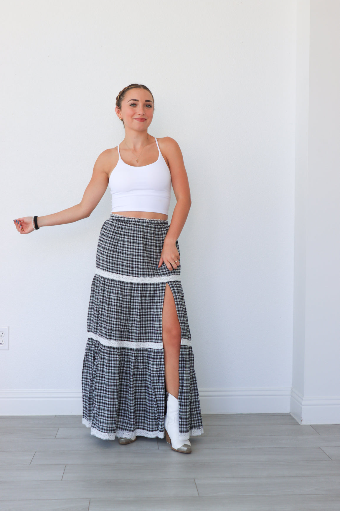 Woman wearing a white sports bra and plaid skirt with a high slit, standing against a white wall.
