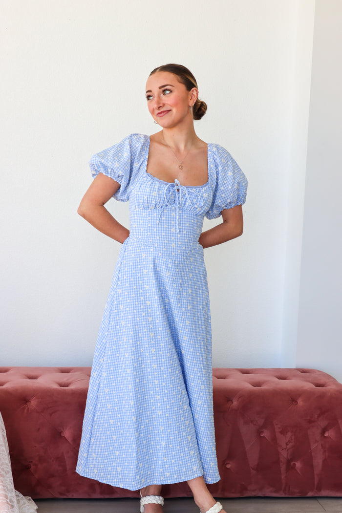 Woman wearing a blue dress with puffed sleeves standing against a white wall.