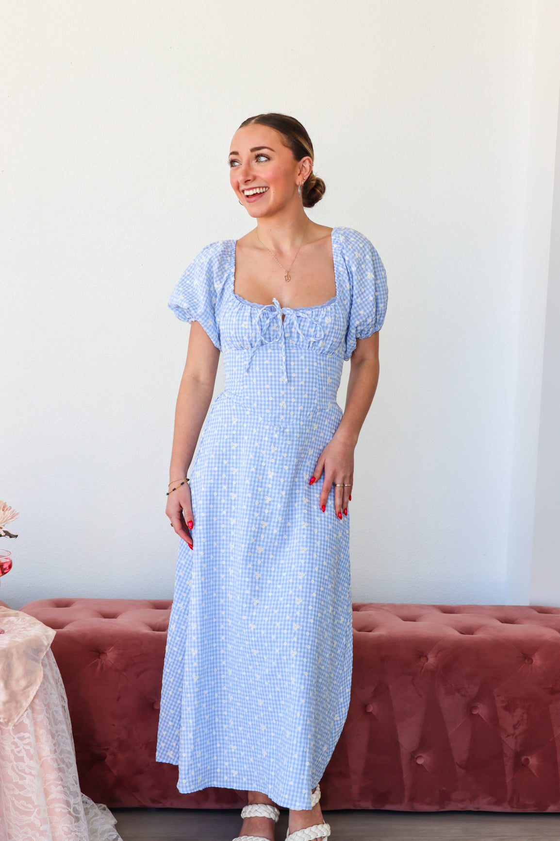 Woman wearing a blue checkered dress standing against a white wall with a red cushioned bench.