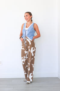 Woman wearing a denim vest over a white shirt and brown camouflage jumpsuit against a white wall.