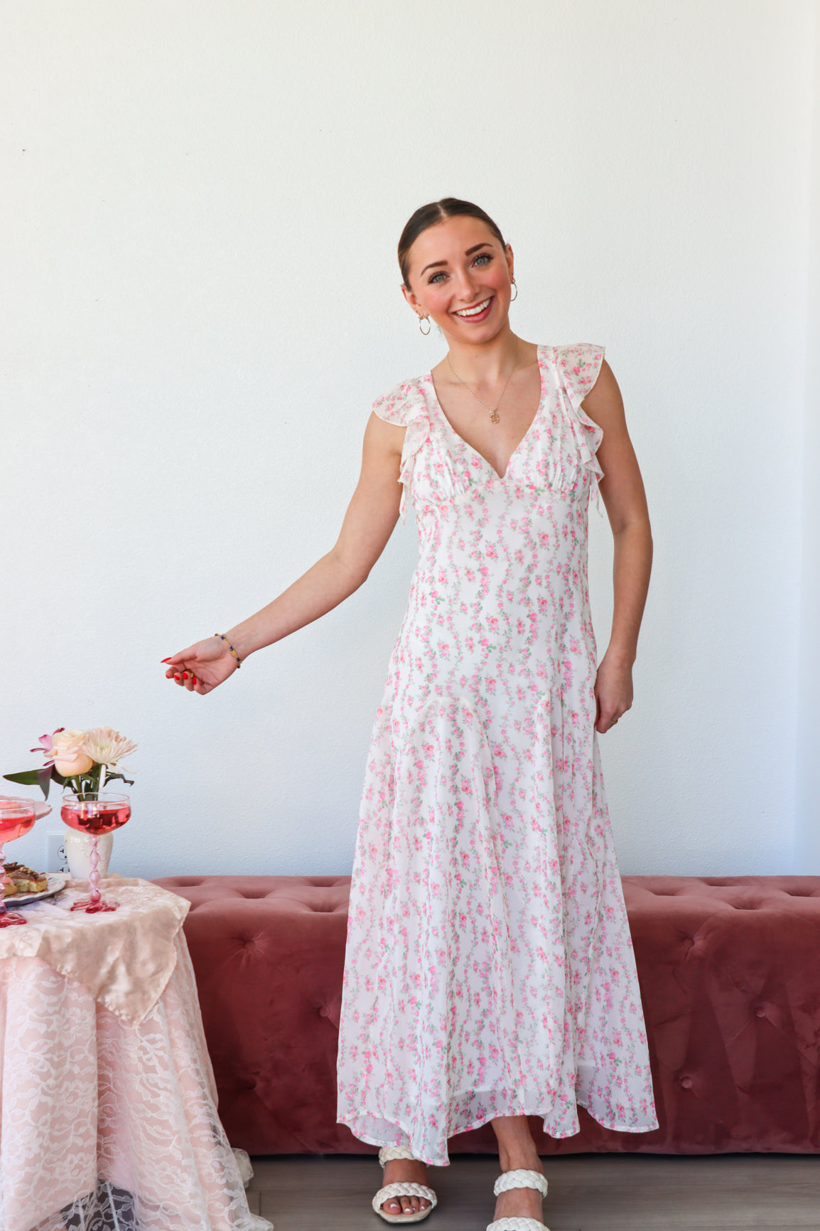 Woman in a floral dress standing next to a table with flowers against a white wall.