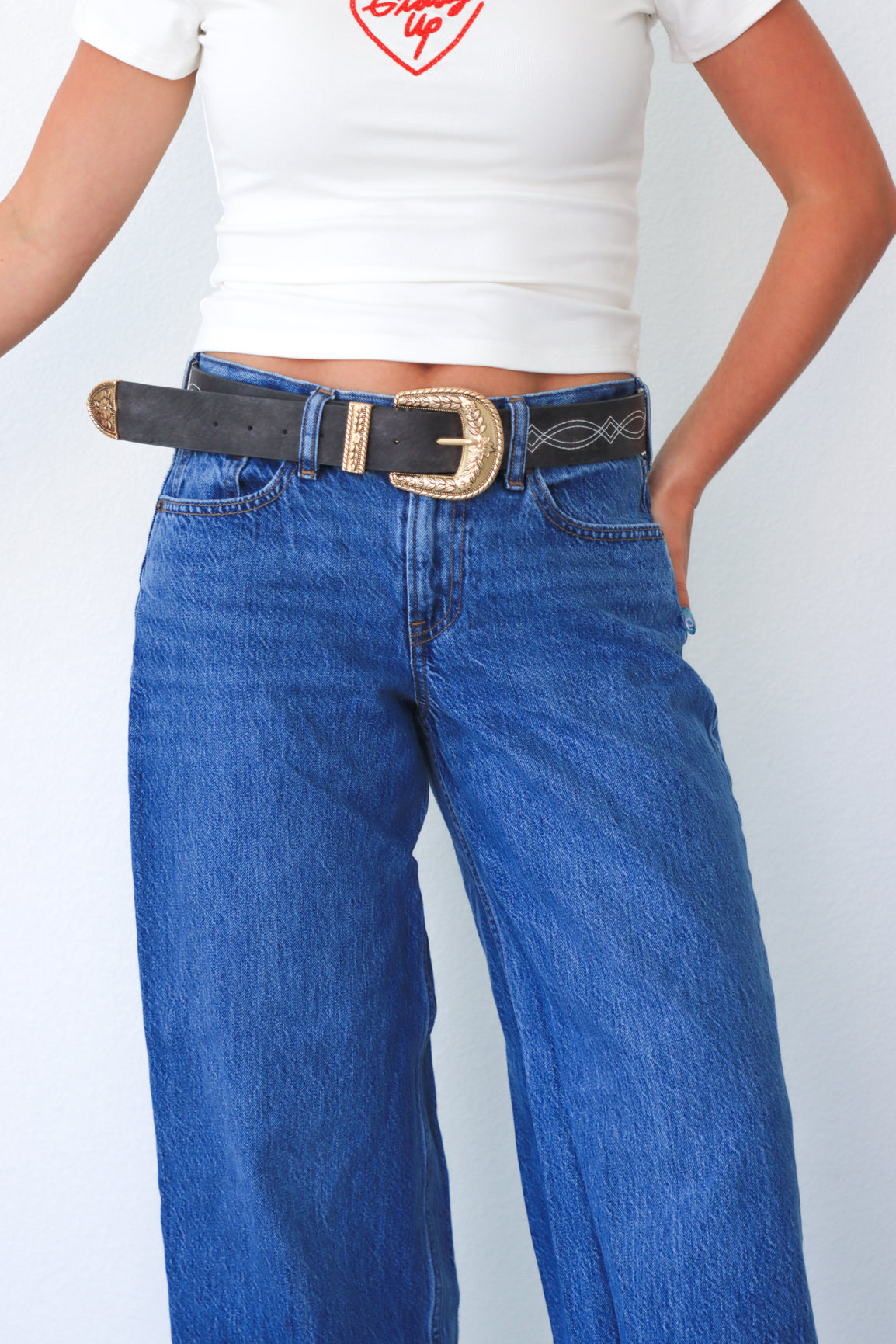 Person wearing blue jeans with a belt and a white top on a plain background