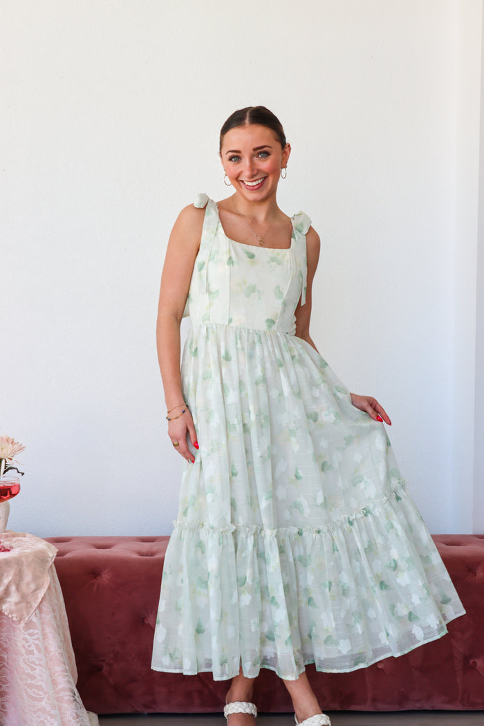 Woman wearing a light green floral dress standing against a white wall.