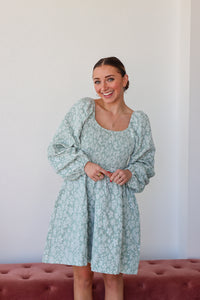 Woman wearing a light blue floral dress standing against a white wall.