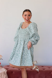 Woman wearing a light blue floral dress standing on a pink couch against a white wall.