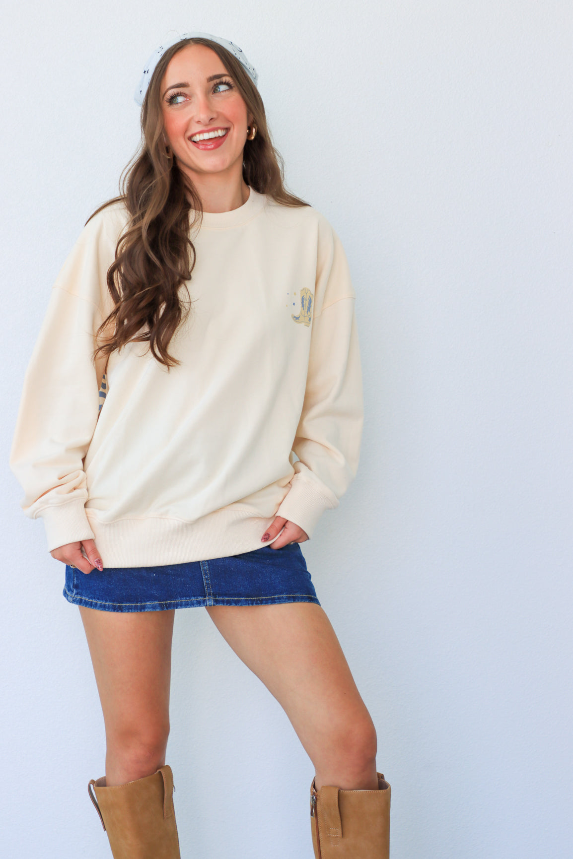 Woman wearing a cream sweatshirt, denim skirt, and brown boots against a white background