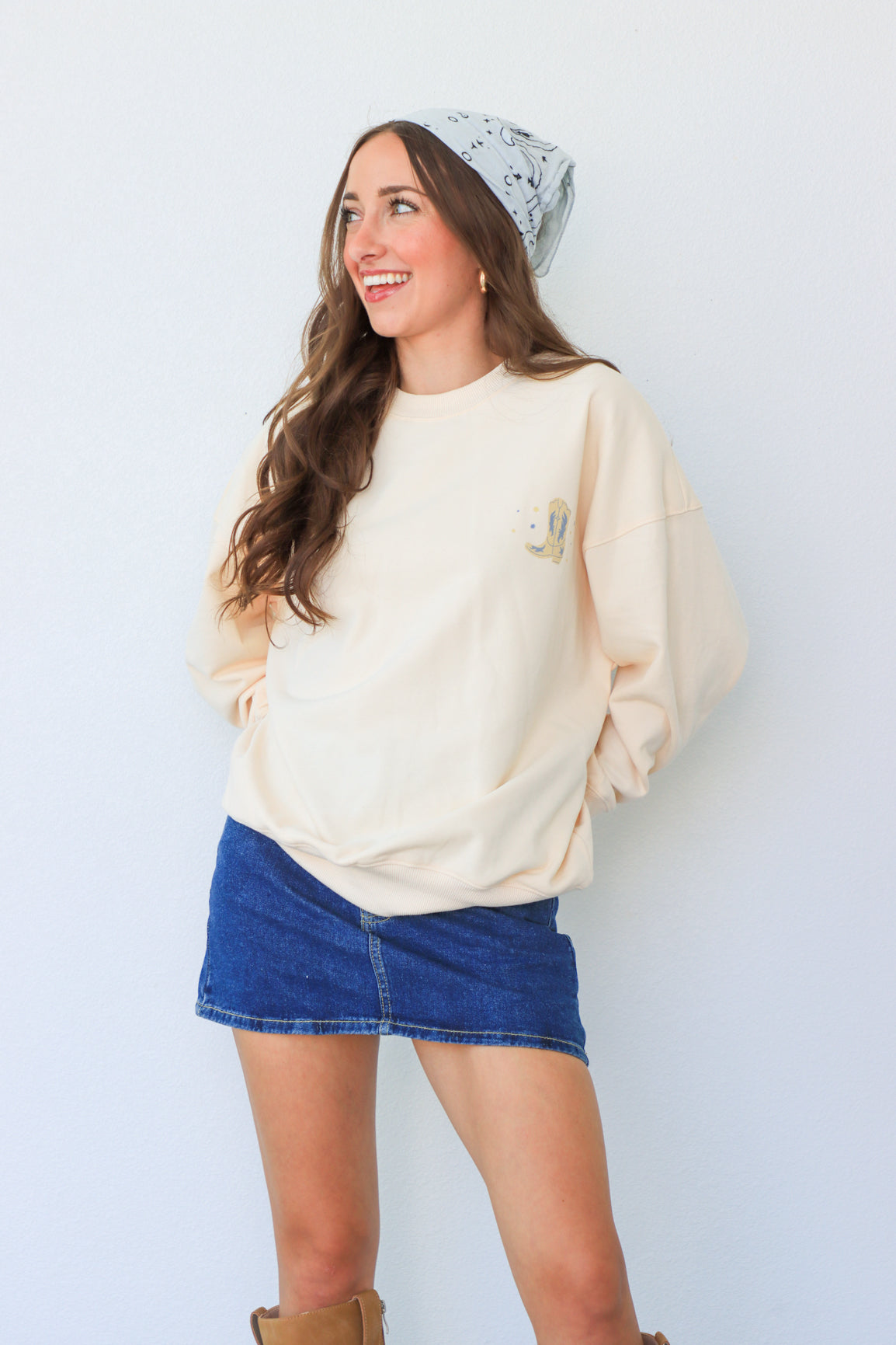Woman wearing a cream sweatshirt with a small design, a denim skirt, and brown boots against a light background