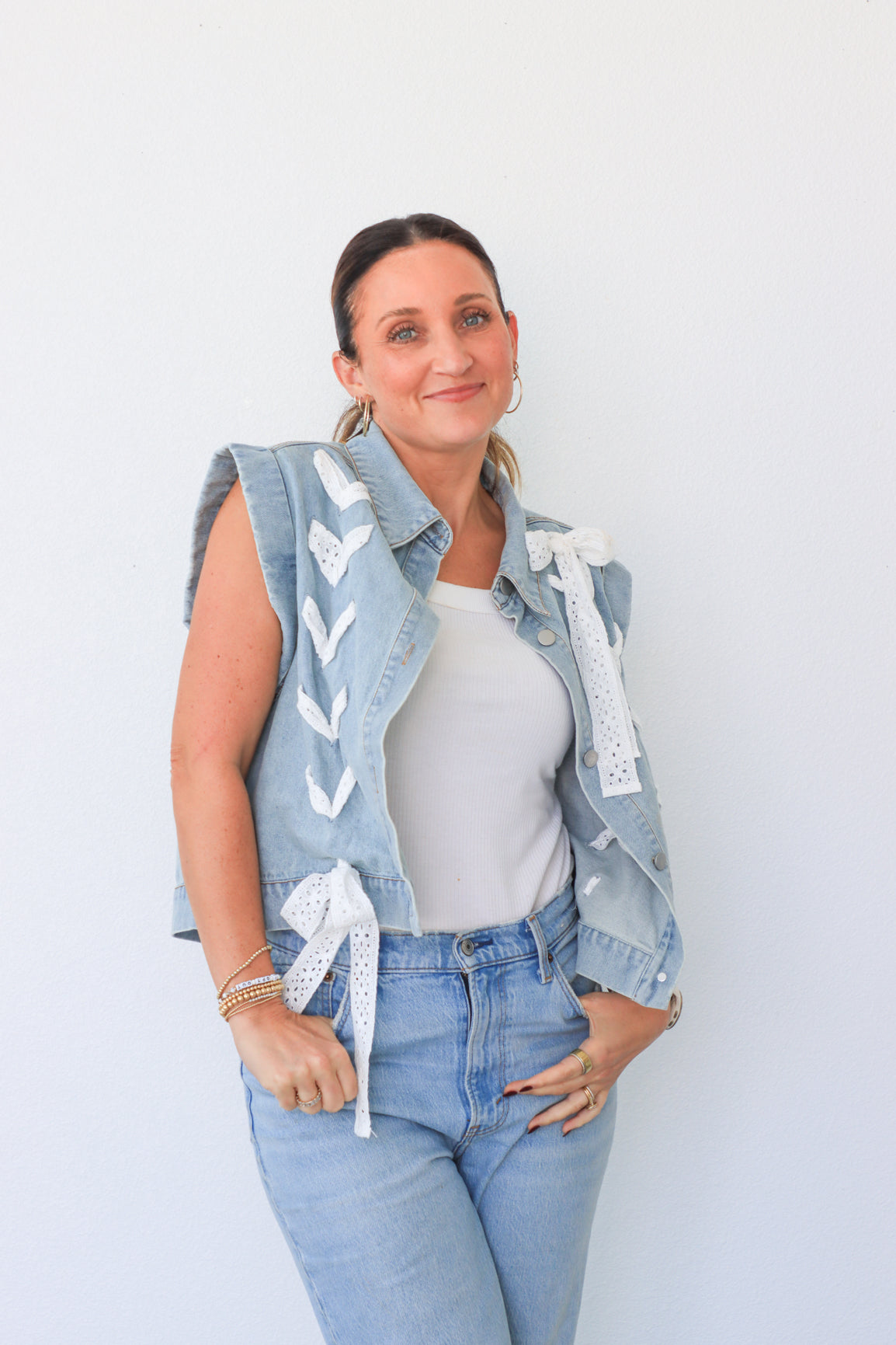 Woman wearing a denim vest with white patterns, white shirt, and blue jeans against a light background