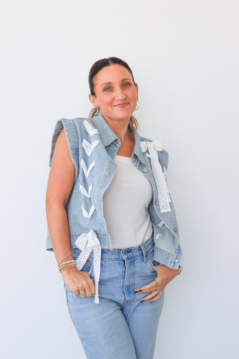 Woman wearing a denim vest with white patterns, white shirt, and blue jeans against a light background