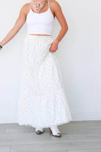 Person wearing a white sleeveless top and long white lace skirt against a plain background