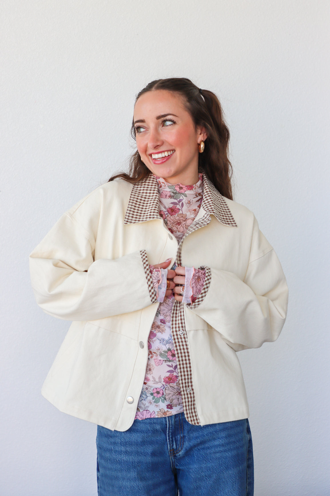 Woman wearing a cream jacket over a floral shirt with a checkered lining, standing against a white background.