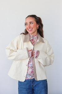 Woman wearing a cream jacket over a floral shirt with a checkered lining, standing against a white background.