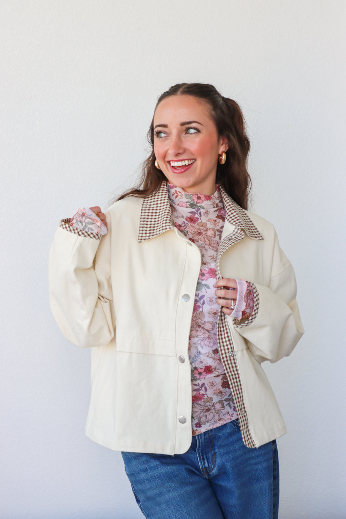 Woman wearing a cream jacket with plaid accents, floral shirt, and jeans against a white background