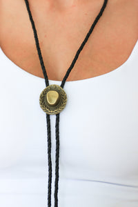 Brass bolo tie on a black cord worn around the neck against a white background