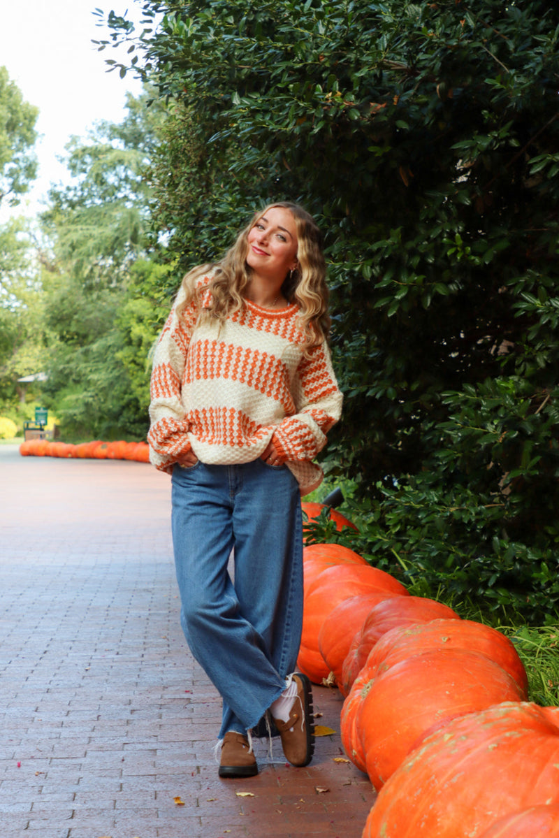 Woman in a striped sweater and blue jeans standing next to pumpkins outdoors.