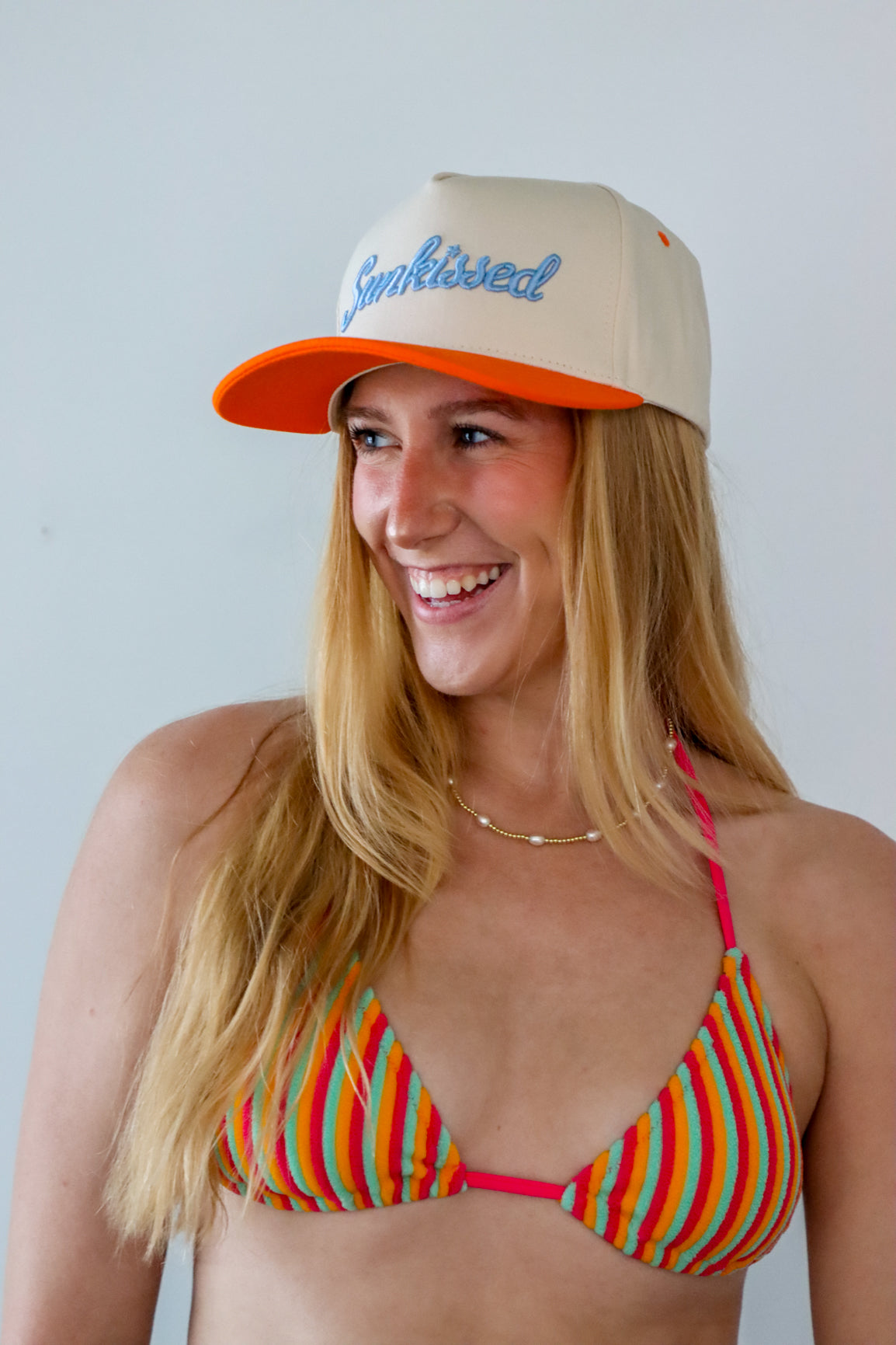Woman wearing a colorful bikini and beige cap with 'Sunkissed' text against a plain background