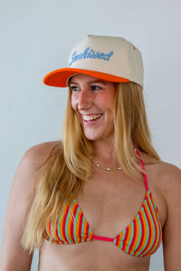 Woman wearing a colorful bikini and beige cap with 'Sunkissed' text against a plain background