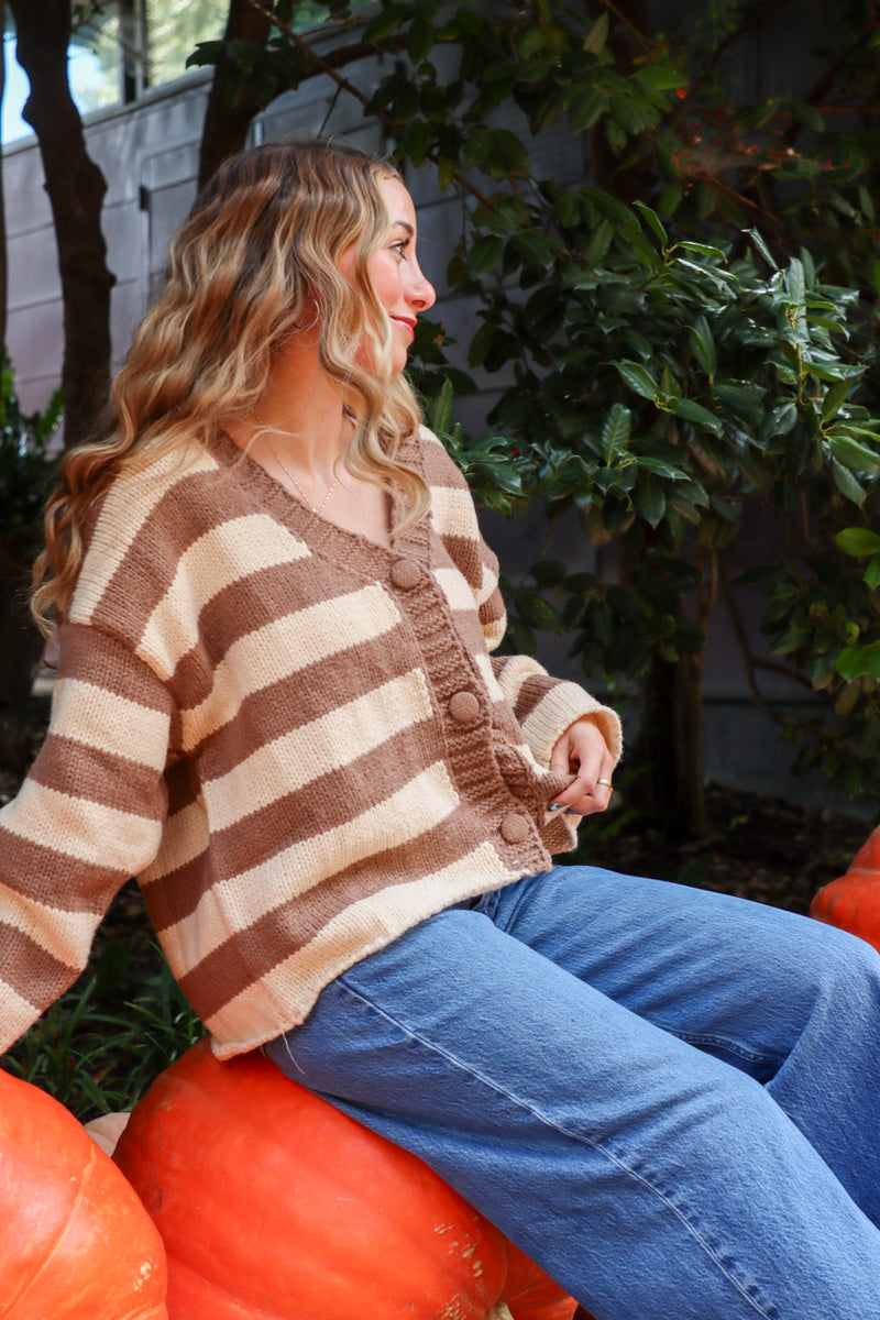 Woman wearing a brown and beige striped sweater sitting on pumpkins outdoors.