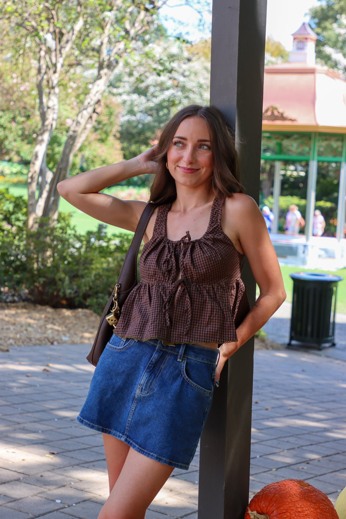 Woman in a brown top and denim skirt standing outdoors with trees and a building in the background.