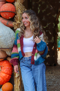 Woman standing among pumpkins wearing a colorful cardigan and jeans.