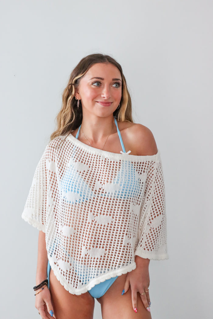 Woman wearing a white crochet cover-up over a blue bikini against a plain background