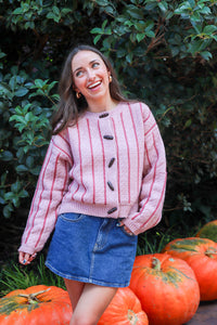 Woman in a pink sweater and denim skirt standing among pumpkins with greenery in the background