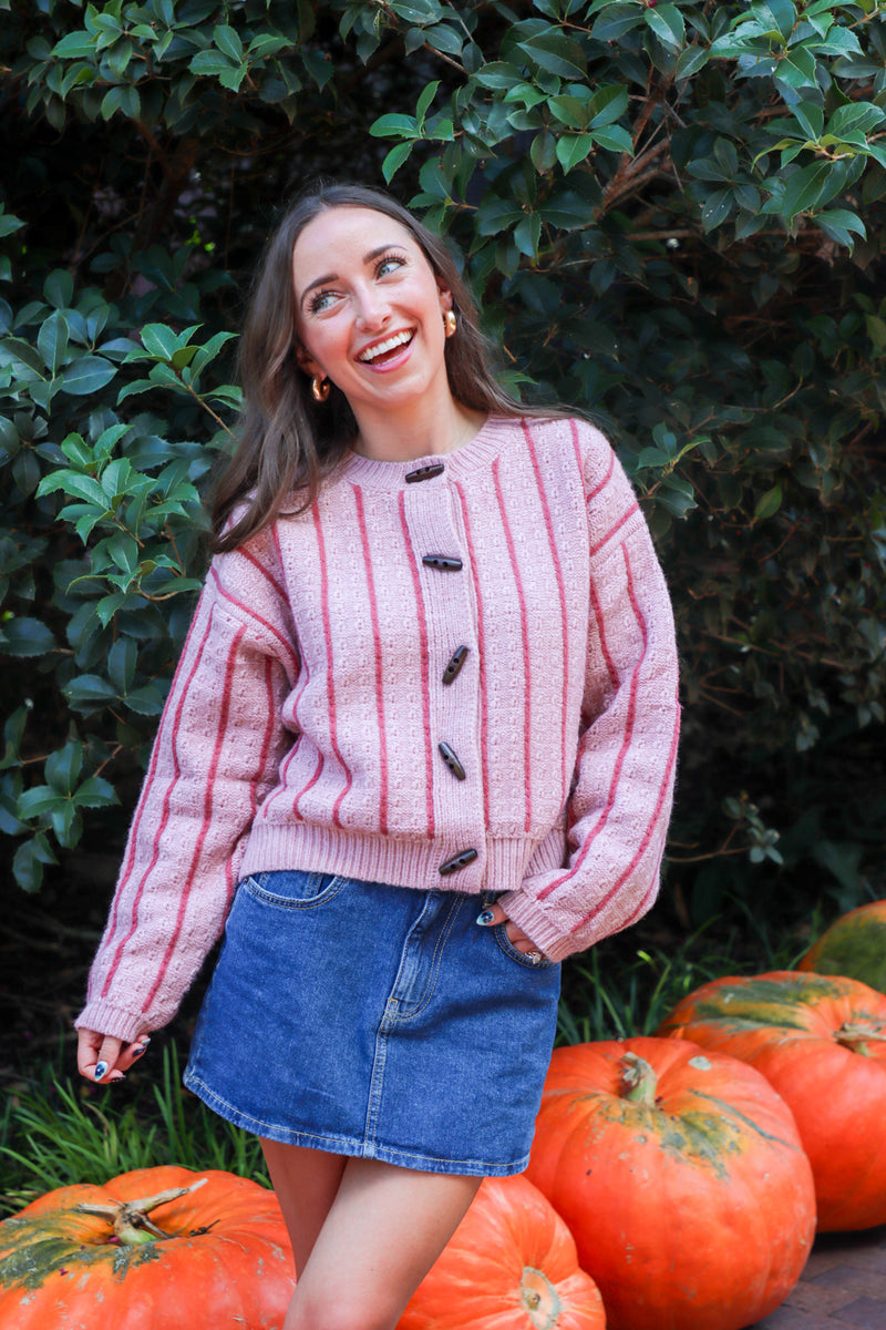 Woman in a pink sweater and denim skirt standing among pumpkins with greenery in the background